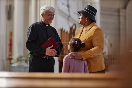 Téléchargez les photos : Prêtre principal discutant cérémonie avec la femme et sa petite-fille pendant qu'ils visitent la vieille église - en image libre de droit