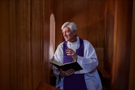Photo for Senior priest reading prayer from Bible while talking to sinner during confession in booth - Royalty Free Image