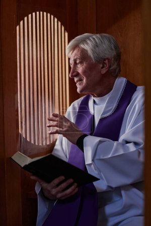Foto de Imagen vertical del sacerdote mayor sentado en cabina con el libro de la Biblia y hablando con el pecador durante la confesión - Imagen libre de derechos
