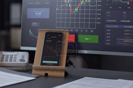 Nahaufnahme eines Smartphones mit Finanzgrafiken auf dem Bildschirm, das auf einem Tisch mit Computern im Büro steht