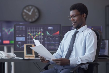 Foto de Comerciante afroamericano examinando documentos financieros mientras está sentado en su lugar de trabajo en la oficina - Imagen libre de derechos