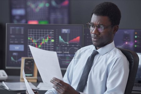 Foto de Hombre de negocios afroamericano en anteojos examinando documento financiero mientras está sentado en la mesa con monitores de computadora con gráficos en segundo plano - Imagen libre de derechos