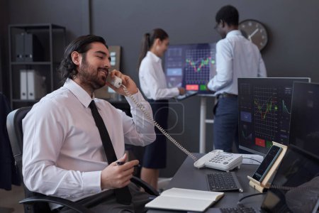 Young broker sitting at his workplace in front of computer monitors and talking to customer by the phone