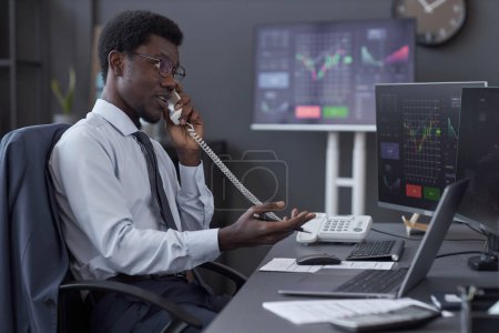 Afroamerikanischer Broker telefoniert, während er am Tisch mit Computern sitzt und mit Kunden über Finanzindikatoren diskutiert