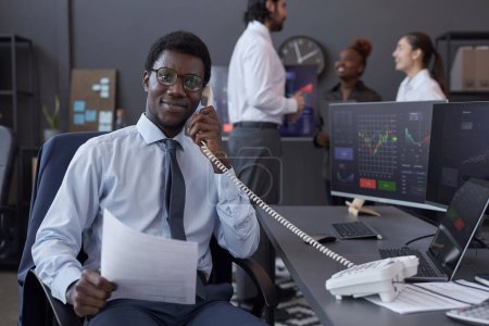Afroamerikanischer Händler blickt in die Kamera, während er mit einem Kunden an seinem Arbeitsplatz über die Lage auf dem Aktienmarkt diskutiert