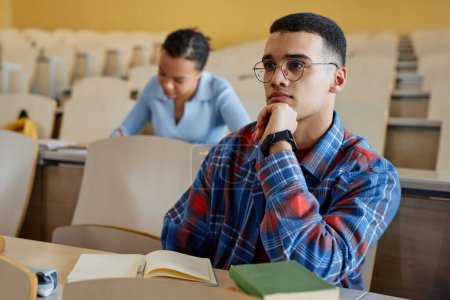 Photo for Teenage boy in eyeglasses sitting at desk and listening to lecturer during lecture in university - Royalty Free Image