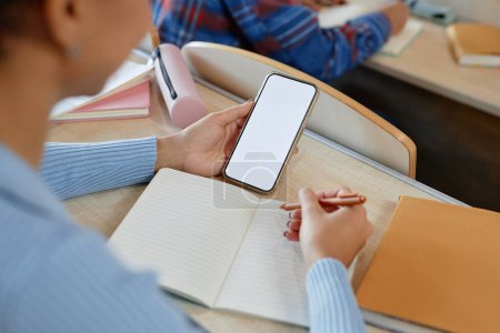 Téléchargez les photos : Vue grand angle de scoolgirl radiant de son smartphone tout en étant assis au bureau avec ordinateur portable à l'examen - en image libre de droit