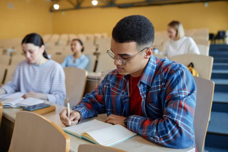 Téléchargez les photos : Adolescent garçon prendre des notes dans notebook tandis que assis à bureau à la conférence avec ses camarades de classe en arrière-plan - en image libre de droit
