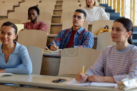 Téléchargez les photos : Groupe d'étudiants assis au bureau et prenant des notes dans des cahiers pendant la conférence à l'université - en image libre de droit