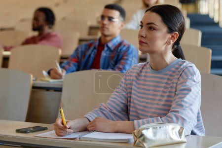 Photo for Young woman sitting at desk and making notes in notebook during lecture with other students in background - Royalty Free Image