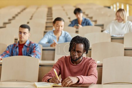 Photo for Group of multiethnic students making notes in notebooks while sitting at desk during lecture - Royalty Free Image