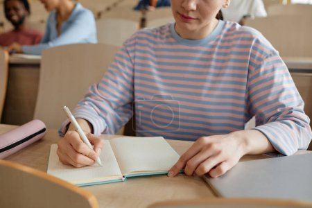 Photo for Close-up of schoolgirl making notes in notebook while sitting at desk during lecture at college - Royalty Free Image