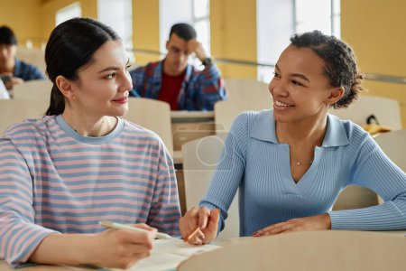Photo for Girl explaining material to her classmate while they sitting together at desk during lecture - Royalty Free Image