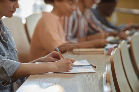 Foto de Primer plano de la colegiala sentada en el escritorio y tomando notas durante la conferencia con sus compañeros sentados en una fila en el escritorio - Imagen libre de derechos
