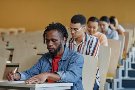 Téléchargez les photos : Groupe d'étudiants assis au bureau dans une rangée pendant l'examen à l'université - en image libre de droit