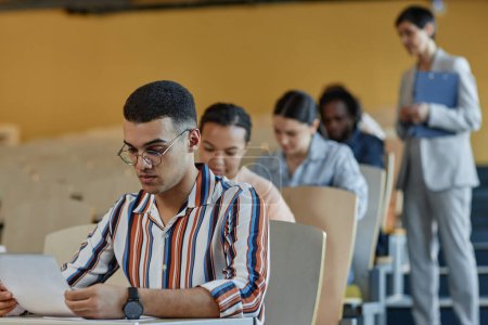 Téléchargez les photos : Groupe d "étudiants qui passent un examen d" écriture assis sur le bureau avec un enseignant contrôlant la situation à l'université - en image libre de droit
