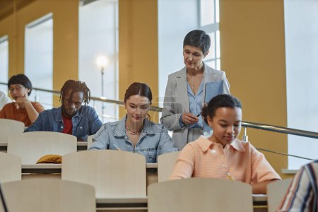 Photo for Mature teacher looking at works of students during exam while they sitting at desk in a row - Royalty Free Image