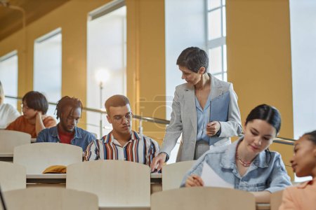 Photo for Teacher walking along the lecture hall and giving hints to students during exam while they sitting at desk in a row - Royalty Free Image