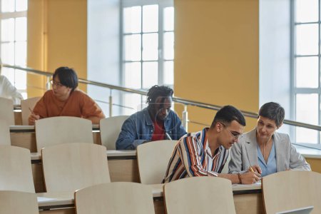 Téléchargez les photos : Professeur mature expliquant matériel à l'étudiant alors qu'ils sont assis au bureau pendant l'examen à l'université - en image libre de droit