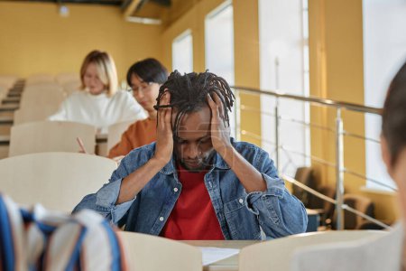 Téléchargez les photos : Afro-Américain écolier tenant sa tête ayant mal à la tête alors qu'il était assis au bureau pendant l'examen - en image libre de droit