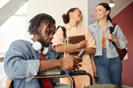 Téléchargez les photos : Étudiant afro-américain handicapé assis sur fauteuil roulant et utilisant un smartphone pendant la pause au collège - en image libre de droit