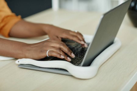Hände einer jungen afroamerikanischen Geschäftsfrau, die am Arbeitsplatz sitzt und auf der Tastatur eines Laptops tippt, während eine weiße Rattenschlange sie umspinnt