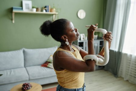 Téléchargez les photos : Blanc rat serpent entrelacement bras de jeune afro-américaine propriétaire tenant animal de compagnie tout en appréciant la thérapie assistée par animal dans le salon - en image libre de droit