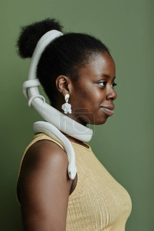 Side view of young black woman with white rat snake enlacing her hair bun standing in front of camera against green background in isolation
