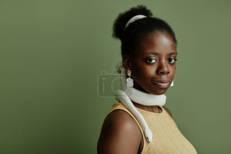 Young African American pretty woman with hair bun and snake enlacing her neck looking at camera while standing on green background