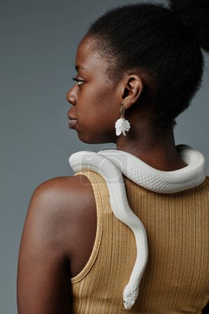 Vista trasera de la joven hermosa mujer afroamericana en pendientes posando con serpiente de rata blanca enlazando su cuello y arrastrándose hacia abajo