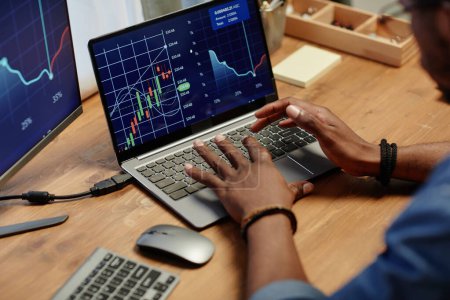 Hände junger afroamerikanischer Broker tippen auf Laptop-Tastatur und analysieren Finanzgrafik auf dem Bildschirm, während sie am Arbeitsplatz sitzen