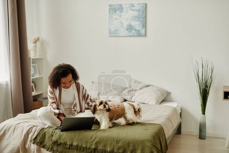 Photo for Cozy portrait of black young woman relaxing at home sitting on bed with cute pet dog and using laptop, copy space - Royalty Free Image