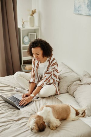Photo for Vertical portrait of black young woman sitting on bed and using laptop enjoying day at home - Royalty Free Image