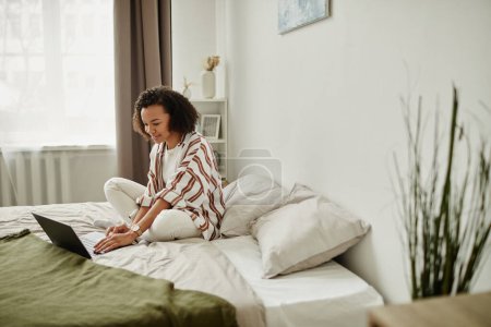 Foto de Retrato de vista lateral de una joven negra usando un portátil en la cama y sonriendo en el interior del hogar mínimo, espacio para copiar - Imagen libre de derechos