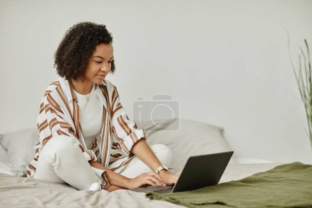 Foto de Retrato mínimo de mujer joven negra usando el ordenador portátil en la cama y sonriendo en el acogedor interior de la casa, espacio de copia - Imagen libre de derechos