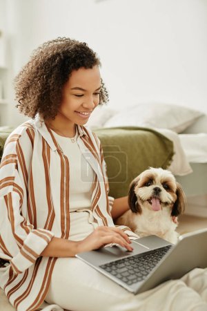 Téléchargez les photos : Portrait vertical de souriante fille noire à l'aide d'un ordinateur portable sur le sol avec petit chien de compagnie - en image libre de droit