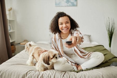 Foto de Retrato de vista frontal de chica negra feliz apuntando control remoto a la cámara mientras disfruta de fin de semana en casa con perro mascota - Imagen libre de derechos