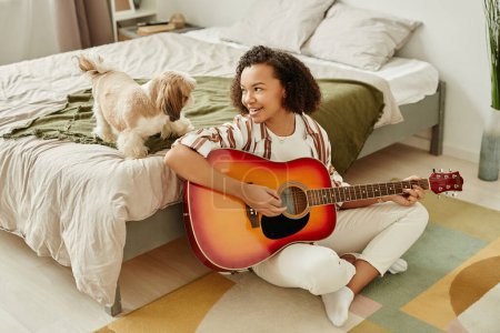 Foto de Retrato franco de una joven negra tocando la guitarra acústica y relajándose en casa con un perrito, espacio para copiar - Imagen libre de derechos