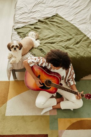 Téléchargez les photos : Vue du dessus à la jeune fille noire jouant de la guitare acoustique et se relaxant à la maison avec petit chien de compagnie - en image libre de droit