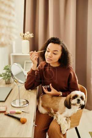 Porträt einer schwarzen jungen Frau beim Make-up am Eitelkeitstisch mit niedlichem kleinen Hund auf dem Schoß
