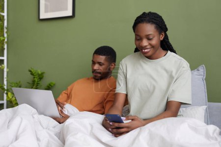 Photo for African American young couple using gadgets while resting in bed during weekends at home - Royalty Free Image