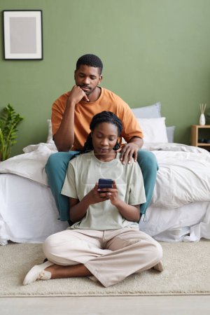 Téléchargez les photos : Jeune couple afro-américain utilisant un smartphone pendant les loisirs dans la chambre à coucher à la maison - en image libre de droit