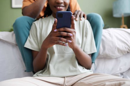 Téléchargez les photos : Gros plan d'une femme afro-américaine textant un message sur son smartphone alors qu'elle était assise dans la chambre avec son petit ami en arrière-plan - en image libre de droit