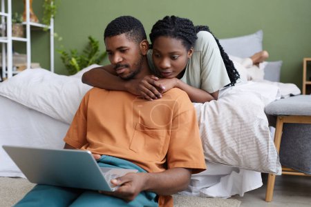 Téléchargez les photos : Afro-Américain charmant couple en utilisant un ordinateur portable le week-end à la maison tout en se reposant dans leur chambre - en image libre de droit