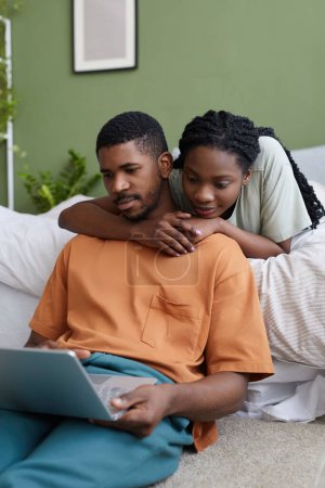 Téléchargez les photos : Image verticale de jeune couple afro-américain regardant un film ensemble sur un ordinateur portable tout en se relaxant au lit dans la chambre - en image libre de droit
