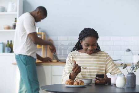 Photo for Smiling African American woman watching funny video on smartphone while having breakfast in the kitchen with her husband in background - Royalty Free Image