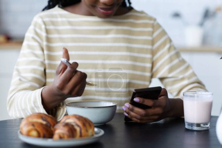 Téléchargez les photos : Gros plan de la femme afro-américaine utilisant un smartphone tout en prenant le petit déjeuner à table dans la cuisine - en image libre de droit