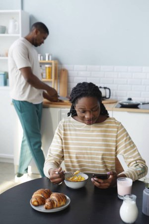 Foto de Imagen vertical de afroamericana usando smartphone y desayunando mientras su marido cocina en segundo plano - Imagen libre de derechos