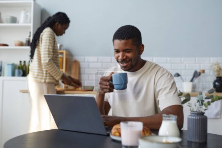 Téléchargez les photos : Afro-Américain utilisant un ordinateur portable tout en prenant le petit déjeuner à table dans la cuisine avec sa femme cuisine en arrière-plan - en image libre de droit