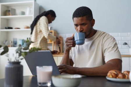 Téléchargez les photos : Afro-américain jeune homme utilisant ordinateur portable pendant le petit déjeuner à table dans la cuisine avec sa petite amie cuisine en arrière-plan - en image libre de droit